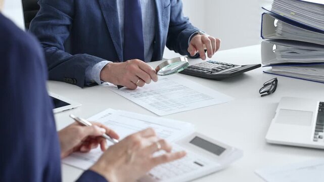 Male financial analysts examining documents, using magnifying glass and calculator while studying corporate data at workplace with a female colleague. Audit and taxes concept