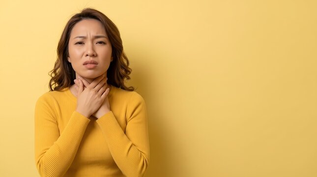 Young Asian woman touching her throat with hands feeling pain and discomfort against yellow background