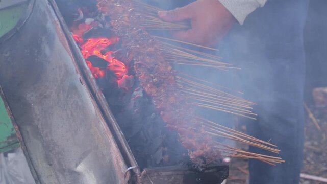 Close-up of satay skewers grilling over burning charcoal with smoke