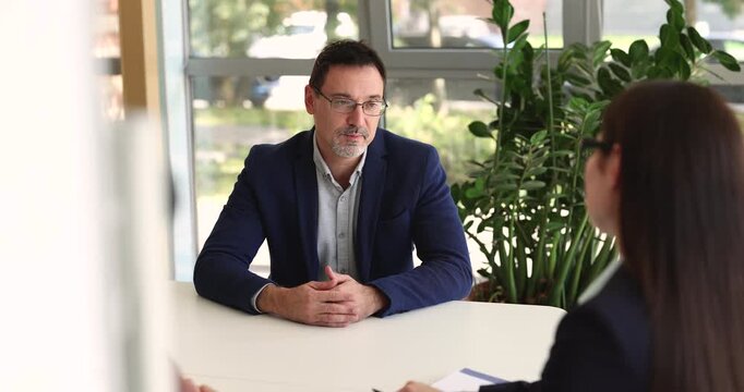 Young woman recruiter speaking with middle aged businessman job applicant during interview discussing decision with unseen colleague and confirming employment agreement with handshake at meeting table