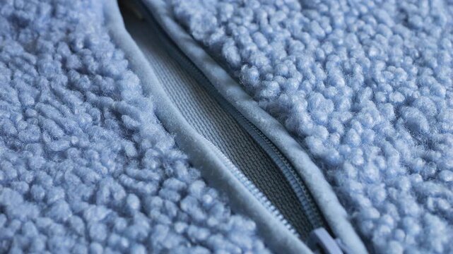 A close-up of a light blue sherpa fleece jacket zipper highlights textured faux fur mimicking curly sheepskin wool, soft boucle fabric warmth, winter outerwear, blue plastic slider, fluffy structure.