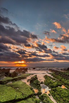 Sunset over Barrika beach with rocky flysch formations and dramatic waves crashing under a golden orange sky, Biscay, Basque Country, Spain