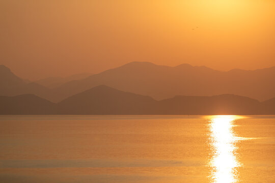A breathtaking golden sunrise casts a warm glow across the calm sea, creating a shimmering reflection on the water. Softly silhouetted hills in the background.