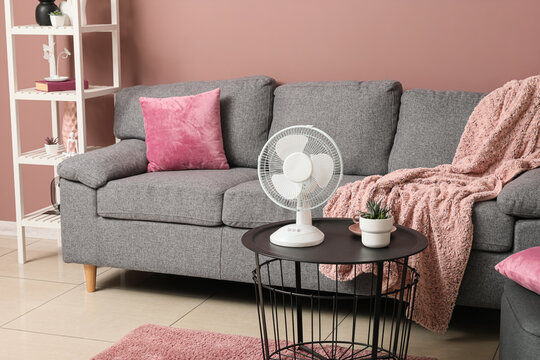 Heatwave relief and home cooling, electric fan on a coffee table in a cozy living room with pink decor and grey sofa for summer comfort