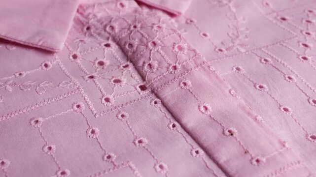 A close-up of a pink cotton women's shirt features delicate eyelet embroidery, batiste material texture, needlework patterns, elegant light color, traditional geometric lace, and sewing art.