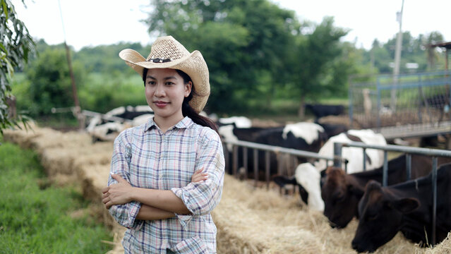Portrait of confident Asian female farmer wearing hat standing with arms crossed at local dairy cow livestock farm.