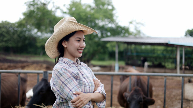 Portrait of confident Asian female farmer wearing hat standing with arms crossed at local dairy cow livestock farm.