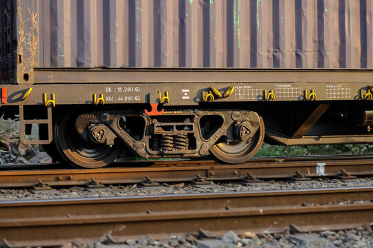 Detailed close-up view of the wheel, bogie, and suspension system of a freight train traveling on railway tracks, featuring industrial metal components, steel rails, gravel ballast, and colorful graff