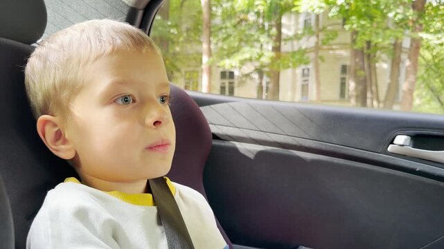 A young boy sits buckled in a car seat and gazes with quiet wonder. He feels happy and calm while riding past green trees on a sunny day.