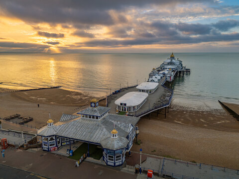 High-angle aerial view of Eastbourne Pier and shingle beach illuminated by dramatic golden sunset light breaking through clouds over the English Channel in East Sussex, England