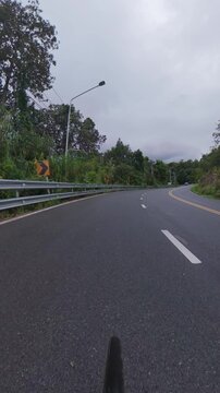 Cyclist POV footage riding along a winding asphalt road through lush green forest, with guardrails, road signs and tropical vegetation on a cloudy day.