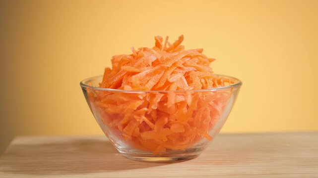 Freshly grated carrots in salad glass bowl against orange background, closeup of vegetables, preparing healthy dieting vegan meal