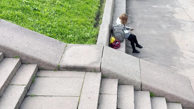 A woman rests with her luggage near granite stairs in Saint Petersburg. She looks calmly at her phone while waiting outdoors. The quiet setting feels peaceful and relaxed.