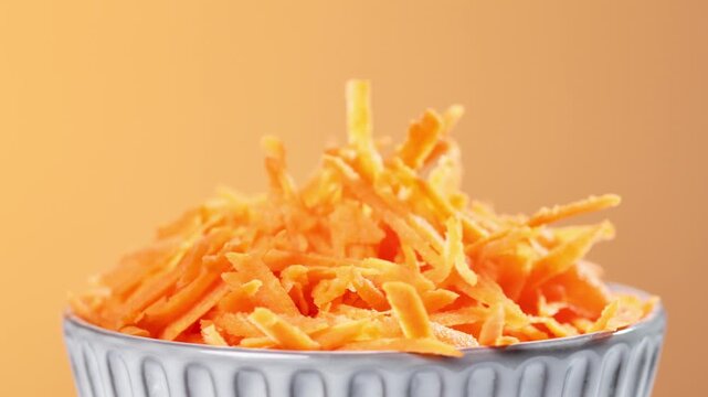 Freshly grated carrots in salad bowl rotate against orange background, closeup of vegetables, preparing healthy dieting vegan meal