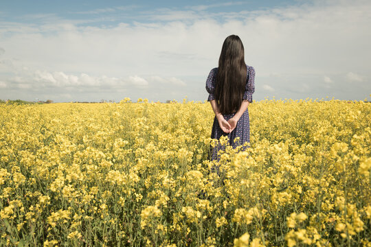fine art portrait of girl in blue dress in field of yellow rapeseed or canola flowers in spring