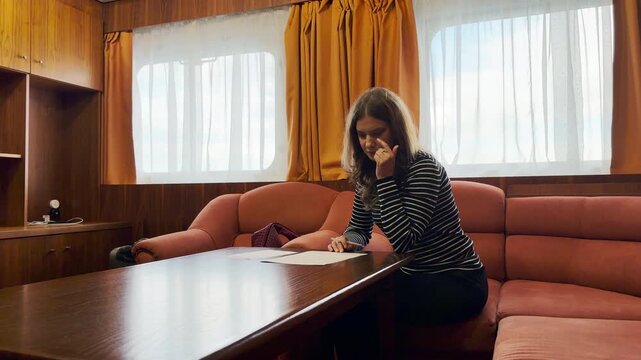 A woman sits thoughtfully reading papers inside a cozy cruise ship suite on the Neva River. She feels calm and relaxed in the warm, peaceful room.