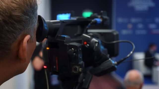 Professional cameraman filming a live conference with video camera. Event broadcasting, live streaming and video production with audience in the background at a business conference.