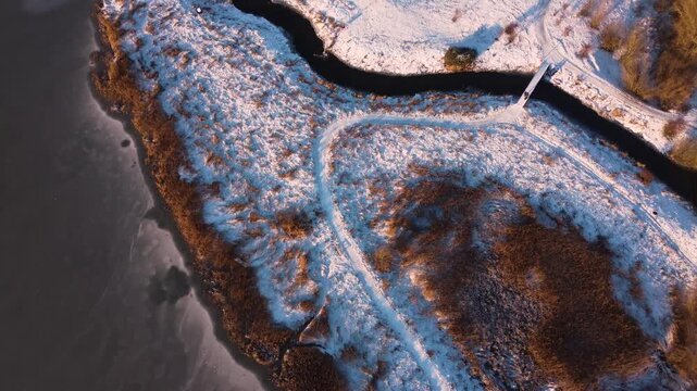 Curving snowy path follows frozen Ega Engso lake wetland shoreline near Aarhus, Denmark, aerial textured backdrop with golden hour sunset light