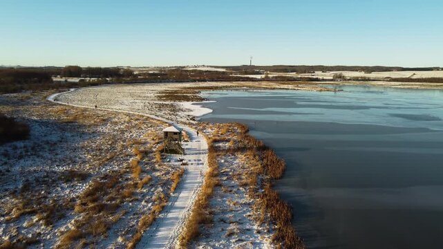 Snow-covered path curves beside frozen Ega Engso lake wetland near Aarhus, Denmark, aerial dolly along sunlit glowing path