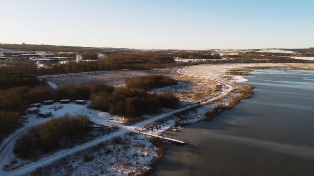 Snowy shoreline and paths beside frozen Ega Engso lake wetland near Aarhus, Denmark, aerial approach to dock with people playing