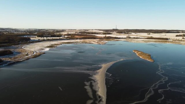 Frozen Ega Engso lake wetland with ice patterns and snowy fields near Aarhus, Denmark, aerial panoramic overview with wispy bands