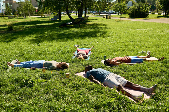 Multiethnic group of young adults lying on mats practicing corpse pose yoga outdoors in park. Visual concept suitable for wellness, mindfulness, and fitness advertising campaigns