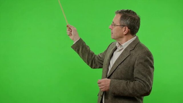 Mature man in glasses and textured jacket gestures with a thin pointer toward the left against a bright green screen background