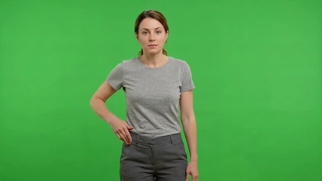 Woman in a gray shirt and dark pants standing against a bright green chroma key background, making different hand gestures and displaying neutral facial expressions in a studio setting