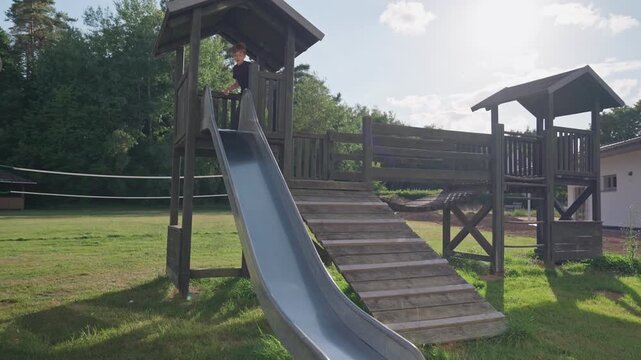 Boy sliding down a metal slide at a wooden playground on a sunny summer day
