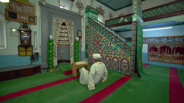 Historic wooden mosque interior with imam, worshippers, carpets, and Islamic prayer setting.