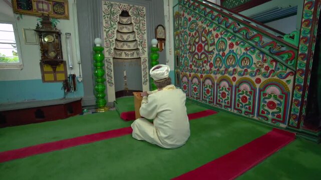 Historic wooden mosque interior with imam, worshippers, carpets, and Islamic prayer setting.