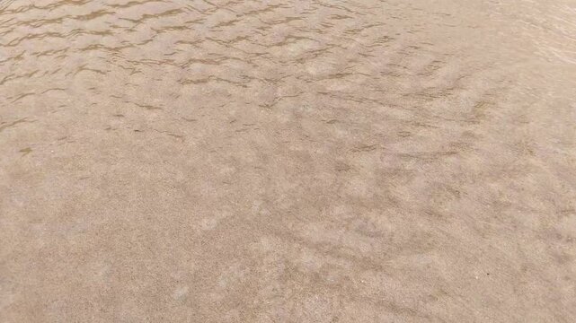 Close-up footage of shallow clear water gently flowing over a sandy riverbed, with sunlight creating rippling caustic patterns across the textured sand surface in a calm natural setting.