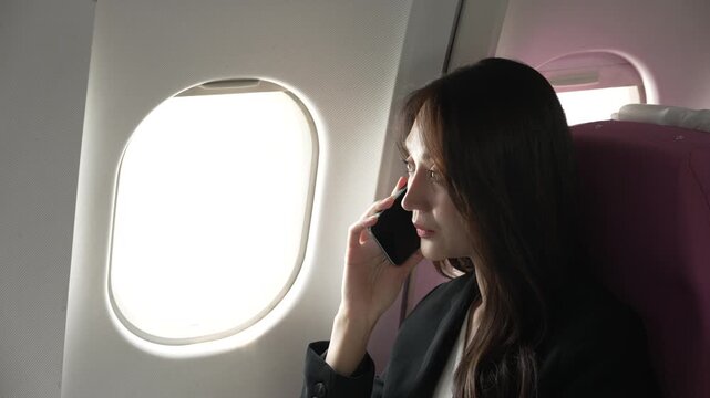 Young woman in a dark jacket sits by an airplane window, looking outside while holding a smartphone during a flight.