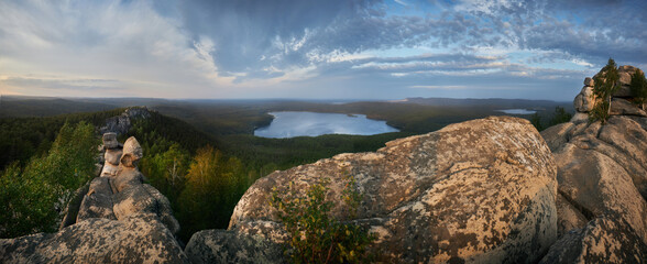 Weathered rocks overlook a forest and distant lake beneath streaked blue clouds in a wide landscape photograph with warm horizon light for outdoor travel themes © angel_nt