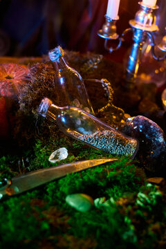 Clear glass bottles rest among moss and crystals beside a pumpkin and burning candles in a vertical fantasy tabletop photograph with colored light for occult themes