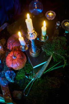 Pumpkins and an ornate knife surround a silver candelabrum on moss and books in an overhead fantasy photograph with glowing candles for autumn and occult design themes