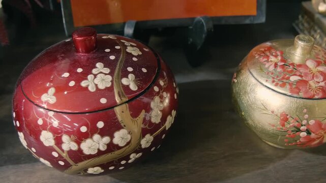 Vietnamese lacquerware trinket boxes crafted from wood and finished with lacquer, incorporating eggshell inlay for detail featuring hand-painted cherry blossom designs stand on a shelf. Closeup