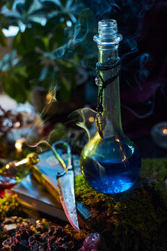 Tall glass bottle of blue liquid stands on books and moss beside an ornate knife in a vertical fantasy photograph with smoke and green foliage for mysterious alchemy themes