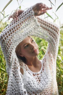 A young woman stands in tall green grass wearing a crochet outfit. She raises her arms and looks thoughtfully towards a bright sky. The setting is natural and summer-like