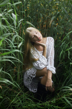 A woman with long hair is sitting in tall green grass. She wears a white outfit and seems to be enjoying the moment. Sunlight filters through the leaves around her