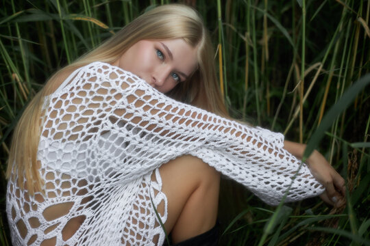 A young woman is sitting among tall green grass wearing a white knit top. She looks directly at the camera while her long blonde hair falls over her shoulder. It is daytime