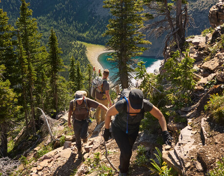 Backpackers hiking up rugged trail on forest hillside above lake, Waterton Lakes National Park