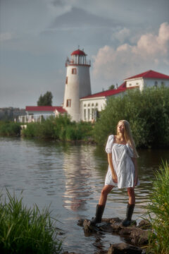 A woman in a white dress poses on rocks in calm water. Behind her is a lighthouse and a building with a red roof. It is a bright day with clouds in the sky