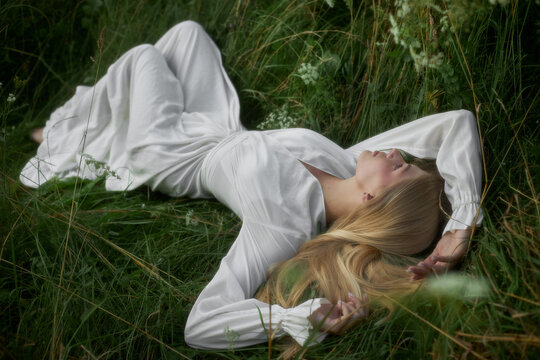 A woman lies on her back in a field of tall grass, wearing a long white dress. Bright sunlight shines down as she relaxes among the flowers. The scene shows a sense of nature