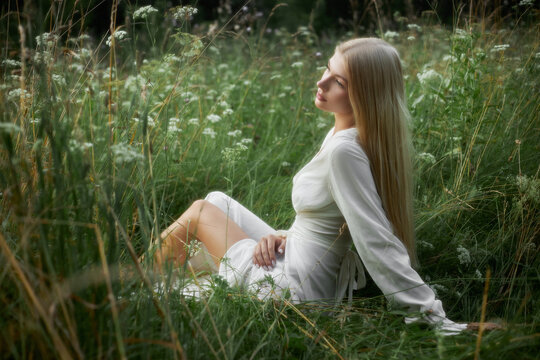 A young woman sits in a field with tall grass and wildflowers. She wears a white dress and has long hair. Sunlight shines down on her as she enjoys the nature around her
