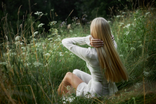 A woman sits in a green field surrounded by wildflowers. She has long hair and wears a light outfit. It looks like a warm day, and she is relaxing in nature