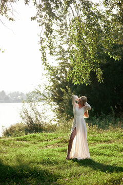 A woman stands near the edge of a lake. She wears a long white dress and plays with her hair. Surrounding her is green grass, trees, and sunlight filtering through the branches
