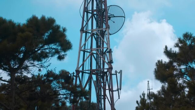 A large telecommunications and broadcasting lattice tower with multiple antennas that sits on the mountain peak in the center against the background of a blue cloudy sky