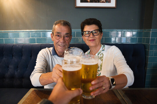 Senior couple and friend clinking beer glasses in a cozy pub booth, smiling and celebrating friendship, leisure and relaxed retirement moments over pints and conversation