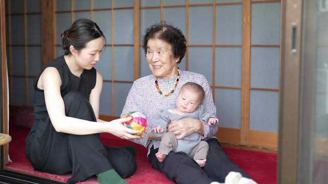 Two adult women sit together with a baby at the threshold of a traditional-style interior, with a colorful ball nearby.